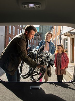 Man loading a folded bike into a car boot, with two people standing nearby on a street.
